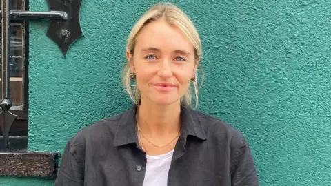 Saffron Billie Croucher, who is blonde and wearing a black shirt, standing against a green wall