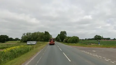 A rural road under a cloudy sky. A signpost on the left reads "Stump Cross Lane." A large red lorry is ahead on the main road, which is surrounded by green fields. 