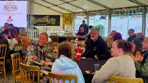 Rach Fosdal People sitting at tables on gold chairs wearing jumpers and coats. They are in a large white marquee with the sides rolled up. A digital screen shows the Mann Made festival logo and there are Bushy's posters to the right of it.