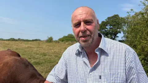Gavin Young is photographed outdoors next a field. He is smiling and looking at the camera. He is wearing a short sleeve white and blue checked shirt.