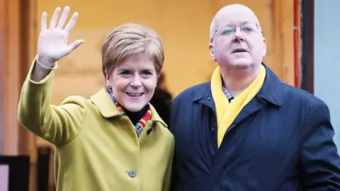 PA Media Nicola Sturgeon, with light-coloured hair, and Peter Murrell, a bald man with glasses, standing side by side in overcoats. Sturgeon is wearing a yellow coat and multi-coloured scarf, while Murrell is in a black coat and yellow scarf. He has his hands in his pockets while Sturgeon has her right hand aloft, waving. 