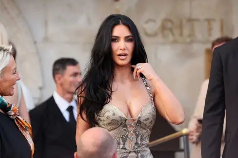 Getty Images Kim Kardashian in Venice, Italy, for the wedding of Jeff Bezos and Lauren Sanchez. She is wearing an animal print dress and has styled her hair long with lose waves. 