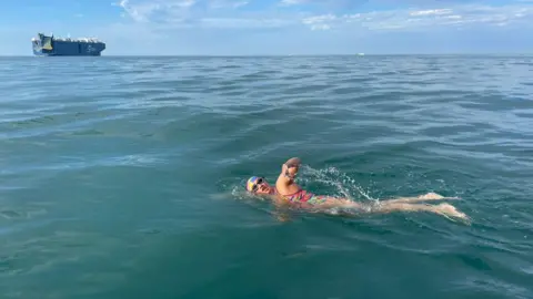 A woman swims in the sea wearing a brightly coloured swimming costume, goggles and a swimming cap. She is swimming front crawl with one arm out the water. In the distance is a large tanker ship and blue sky.