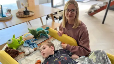 Sarah, who is blonde and has a dusty pink sweatshirt on, is next to Toby's bed, he is lying down surrounded by cuddly toys