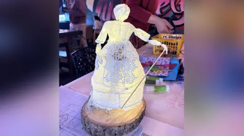 A maquette of an illuminated puppet which is being created to be called the Spirit of Fleetwood. It is standing on a piece of wood on a desk with sketchings of the figure. 