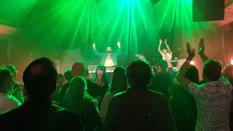 The stage is bathed in green light as Flamingods perform. The camera shows two men on stage, with their arms aloft encouraging the audience to clap along.
The camera is behind the audience and shows their back views, apart from one woman who has turned round.