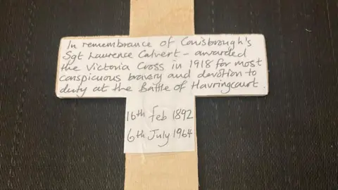 John Healey A close-up of the cross planted - a piece of paper is taped to it. A handwritten message on the paper reads: "In remembrance of Conisbrough's Sgt Laurence Calvert - awarded the Victoria Cross in 1918 for most conspicuous bravery and devotion to duty at the Battle of Havrincourt. 16th Feb 1892. 6th July 1964."