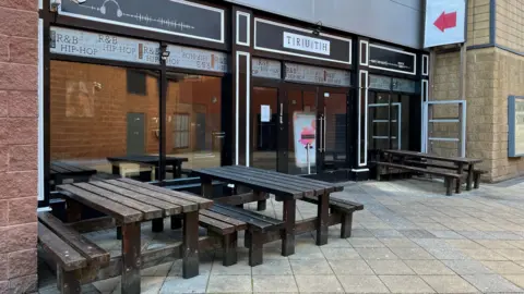 BBC The entrance of Truth nightclub. Glass doors with black panes lead into the venue, with club logo above. There are wooden benches and tables at a seating area outside.