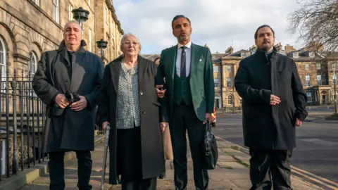 PA Media Emma's mother Margaret Caldwell, uncle Jim Coyle, nephew Stewart McGrory and lawyer Aamer Anwar address the media outside Bute House on a sunny day - all three family members are wearing dark coats while Ms Caldwell is holding a walking stick. Mr Anwar is wearing a green suit and is the person speaking.