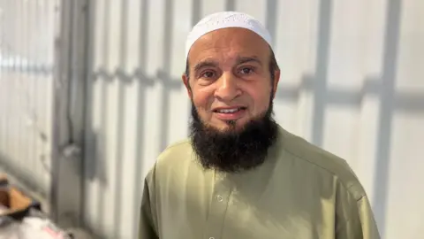 Elizabeth Baines / BBC A man in a green shirt smiles. He has a beard and wears a white cap. Behind him, a metal structure is blurred