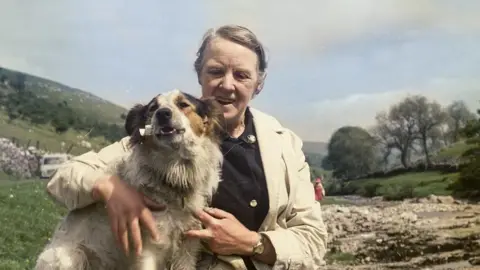 A woman with grey hair on a moor wearing a mac and a black cardigan hugs a white border collie dog.