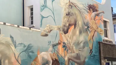 Cheltenham Borough Council A mural showing a white horse with women dancing around it on the side of a building
