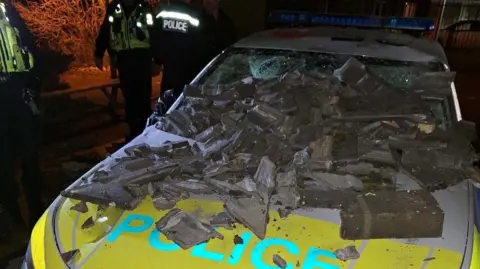 A police car with smashed ceramic tiles on its front windscreen and bonnet. The windscreen has been smashed by the tiles. Uniformed offices stand nearby.
