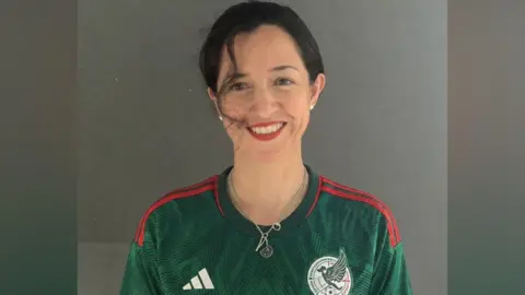 Catalina Gomez A woman with tied-up black hair smiling at the camera. She is wearing a green Mexico football shirt.