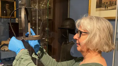 Leeds Museums and Galleries A woman with white hair holds an iron device. She is wearing blue gloves, a green top and black rimmed glasses.