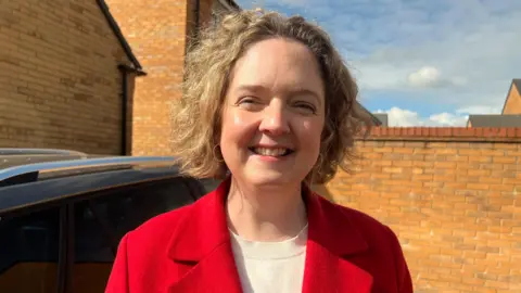 BBC Anna Sabine is smiling at the camera. She is wearing a beige jumper and a red jacket. There is a car parked to the left of the image and new build houses are behind her.