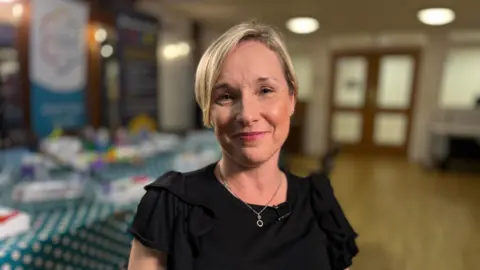 Martin Giles/BBC A smiling Amanda Orange looks directly at the camera. She is standing in a hall, and is wearing a black blouse and silver necklace. 