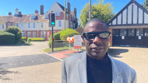 A man with short hair is wearing a grey jacket. A pelican crossing and two buildings are behind him.
