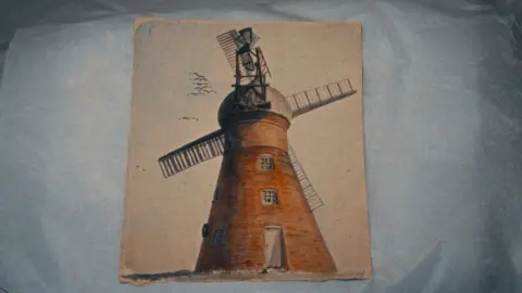 Emily Andersen A film still showing a painting of a red-brick windmill with four slatted sails. The mill has two square windows and a white door. Birds are flying across a hazy sky.