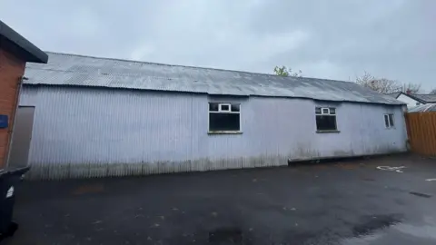The image shows a long single-level building made from corrugated metal. There are three windows on the side and a tarmac carpark in front. On one side is a wooden fence, on the other is the corner of a red brick building. 