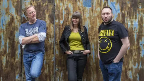 Two men and a woman are standing in front of a wall that is metal and rusted. The man on the left has short blonde hair and a white goatee. The woman has brown shoulder length hair. The man on the right has short black hair with black facial hair. He is wearing a black and yellow Nirvana t-shirt