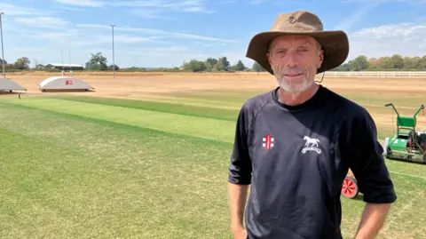 Richard Mosdell stands in front of a watered central square on a cricket pitch while an arid dry and un-watered area can be seen in the background. he is wearing a dark long-sleeved top and has a wide-brimmed dark green sunhat on his head