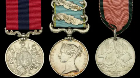 Three military medals displayed side by side. The left medal has a silver circular disc with an intricate design featuring a crown, flags, and weaponry, attached to a ribbon with vertical stripes in dark blue and crimson. The centre medal shows a silver disc with a profile of a monarch, attached to a light blue ribbon adorned with four silver clasps. The right medal features a silver disc with a design of crossed flags and weapons, attached to a ribbon with alternating salmon-pink and green stripes.
