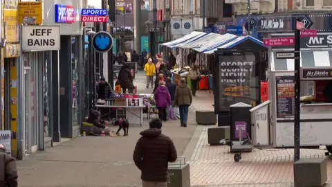 A shot of a Walsall town centre street, showing shoppers, a market and shops. A beggar sits on the floor in a sleeping bag with a black dog next to him.