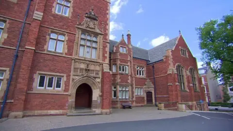 Red brick exterior of ornate Methody college, with light stone details around windows, blue sky and some green trees visible in shot