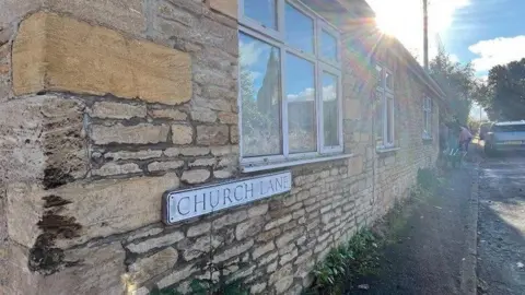 Warmington Almshouses Charitable Trust Side exterior of a historic-looking stone house with dated windows with white frames.