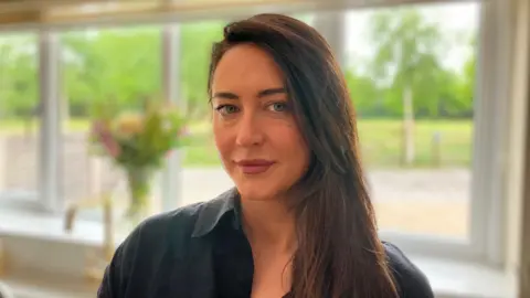 A woman with long dark hair, swept over one shoulder. She is wearing a dark top, and is in front of a window with a field behind her