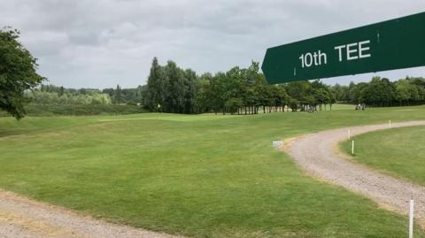 Waterstock Golf Course, with a sign in the foreground pointing to '10th Tee'. A golfing green with a flag is in the background. Golfers are in the far distance, with trees behind them.