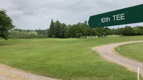 BBC Waterstock Golf Course, with a sign in the foreground pointing to '10th Tee'. A golfing green with a flag is in the background. Golfers are in the far distance, with trees behind them.