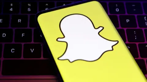 A smartphone showing Snapchat's logo, a white, faceless ghost figure on a yellow background, in front of a keybaord.