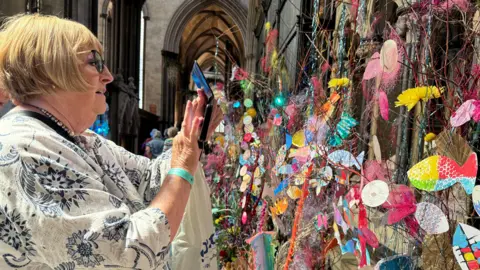 A lady wearing glasses and a white top taking a picture of a very colourful display made with arts and crafts on a wall.