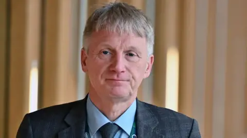 Getty Images Ivan McKee, who has short grey hair, looks towards the camera. He is in front of wooden panelling and is wearing a grey suit and tie with a blue shirt. 