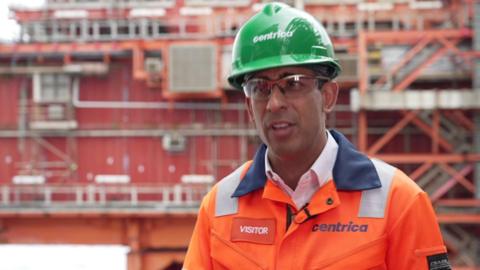 Rishi Sunak stands near a building site wearing a green hard hat and orange high-visibility jacket.
