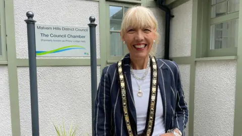 Malvern Hills District Council Beverley Nielsen pictured beside a sign for Malvern Hills District Council. She has blonde hair which has been tied back and is wearing a navy blue jacket with white stripes. She is also wearing gold jewellery and a ceremonial gold chain around her neck.