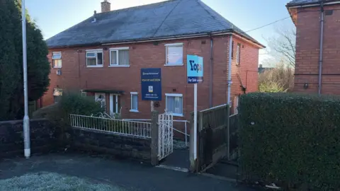 Two neighbouring houses both display sales boards. It is a frosty, clear day, there is ice on the ground, and the houses have a wall and a hedge in front of them. There are gates to the paths leading to the front doors.