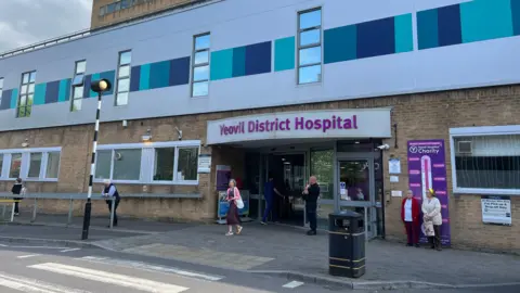 The entrance to a hospital is pictured with a sign saying Yeovil District Hospital. Outside is a zebra crossing on a road.