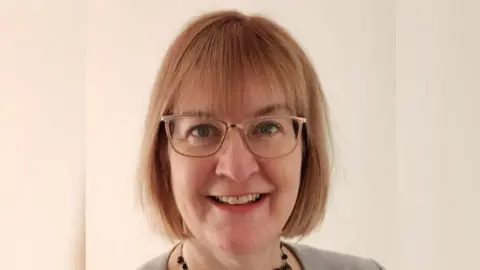 Headshot of Jo Walker, a middle-aged woman with a strawberry blonde, fringed bob and light rimmed glasses, smiles. She is wearing a black beaded necklace and the shoulders of her grey top can be seen. She has blue eyes and is standing against a plain, cream-coloured background.