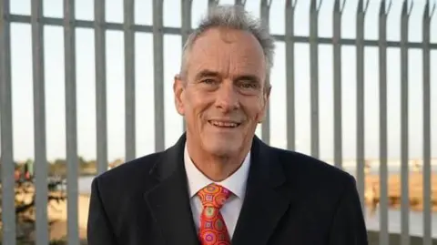Shaun Whitmore/BBC Giles Membrey is standing outside in front of a metal fence. He is looking directly at the camera and smiling and is wearing a black coat with a white shirt underneath and a red patterned tie.