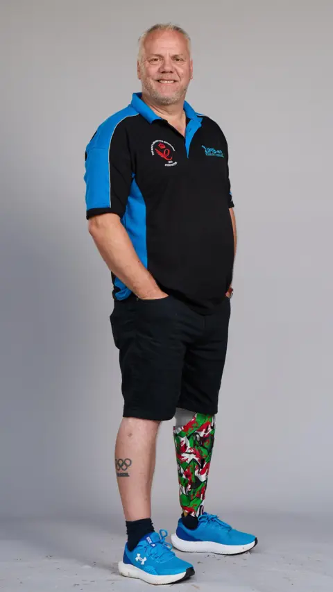 Department of Business and Trade Mark poses for a full length photo in a studio. He has close-cropped grey hair and a stubbly beard. He's wearing a black and blue T-shirt with his company logo, black shorts and blue trainers. He has the Olympic rings tattooed on his right leg, and his left prosthetic leg has a cover attached to it mimicking the shape of a lower leg. The cover is brightly patterned with the red, green and white colours of the Welsh flag.