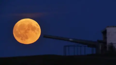 PA Media An image of a Buck Moon (which looks like a golden yellow) to the left of a canon coming from the right. 