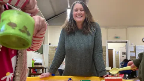 Simon Thake/BBC A smiley woman with long dark hair wearing a green jumper stands infront of a low children's table where small children's hands can be seen playing with buckets and spades