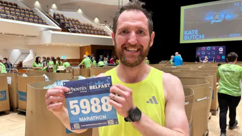 Aaron Beggs, with short brown hair and a brown beard, wearing a sleeveless yellow sports top. He is holding a blue page with the number 5988 written in white. 