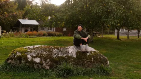 Jesse Morrow Brian Fennell is sitting on a  large rock in a garden with trees concealing a house. He is wearing a green hoodie, dark trousers and a dark beanie hat.