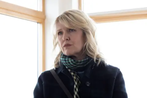 BBC/Silverprint Films/Jamie Simpson Ashley Jensen looks off to the right with concern on her face. Bright light floods in through a corner window behind her. She is wearing her dark coat and patterned scarf