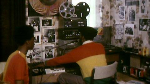 A man and woman wearing bright jumpers sit in a DIY radio studio