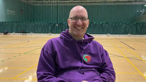 Adam Simmons, in a purple hoodie with a red hawk logo on it. He is wearing glassed and is smiling directly at the camera. Behind him is the rugby court. There is a green mesh curtain in the background.